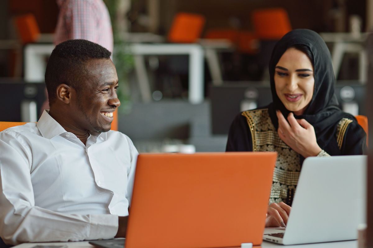 Two people smiling while working on laptops, one orange and one white, in an office with orange chairs and blurred background.