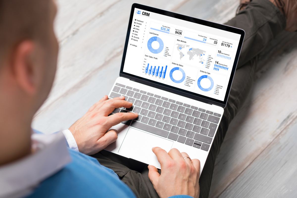 Person using a laptop displaying CRM data with charts and graphs. The user sits on a wooden floor, wearing a blue shirt. Mood is focused.