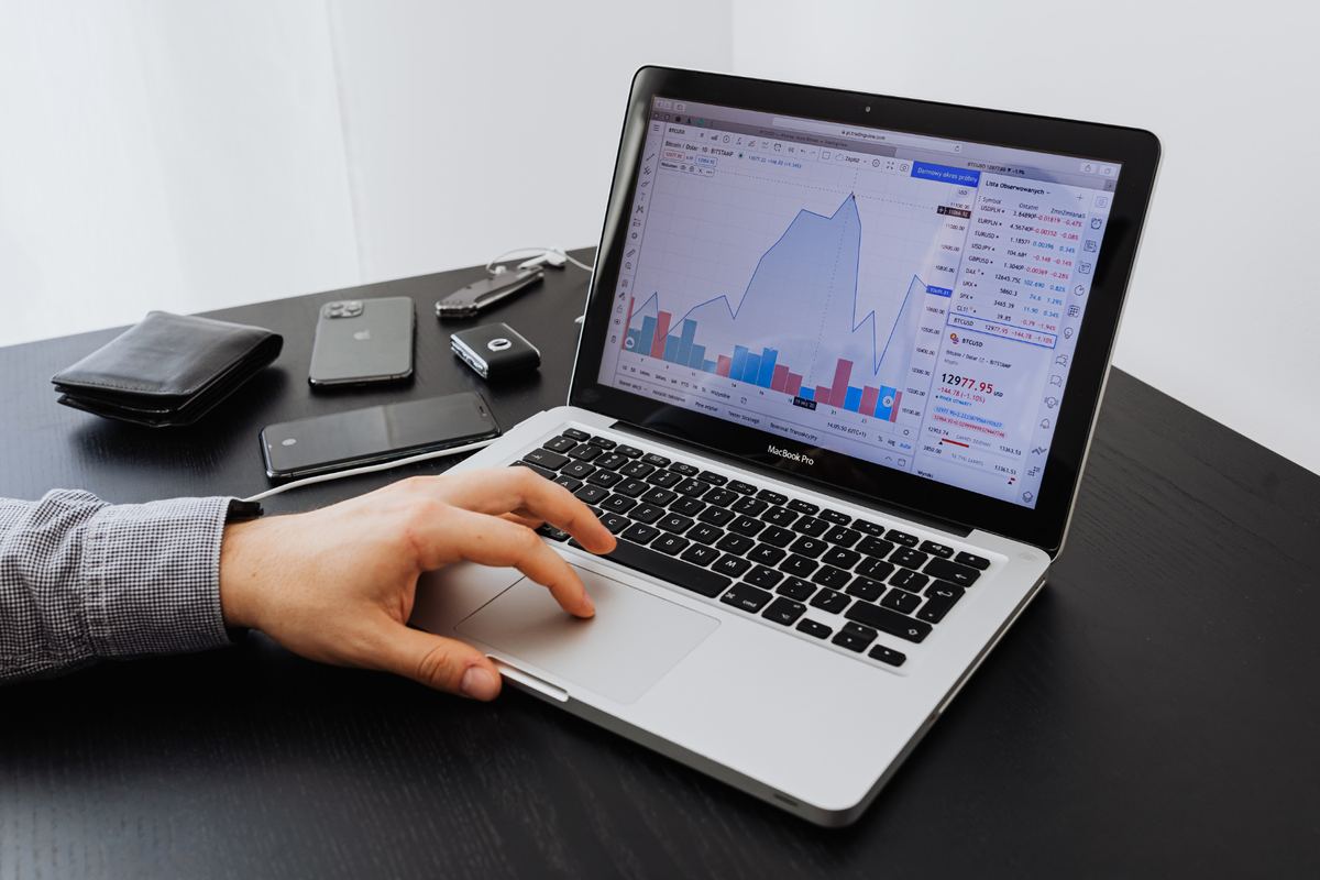 Hand interacting with a laptop displaying financial graphs. Black table with a phone, wallet, and keys nearby. Bright, professional setting.