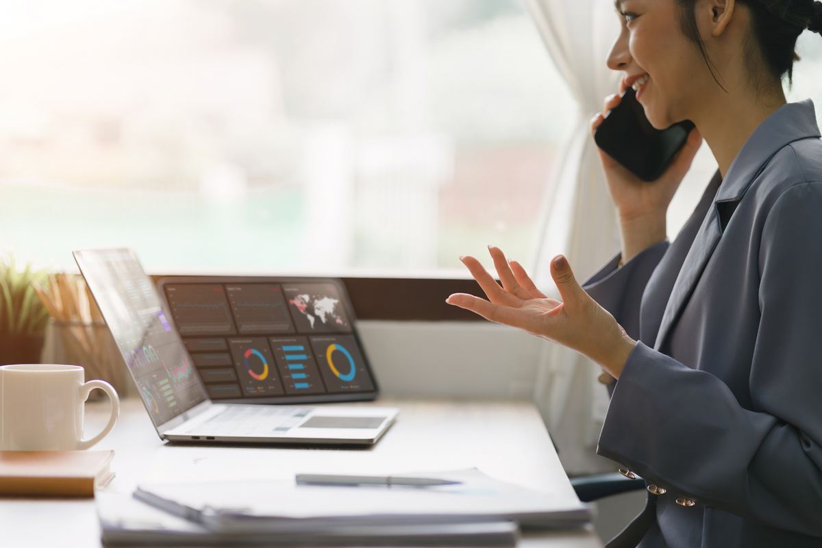 Woman in a blue suit on a phone call, gesturing near a desk with a laptop displaying graphs. Bright, airy office setting.