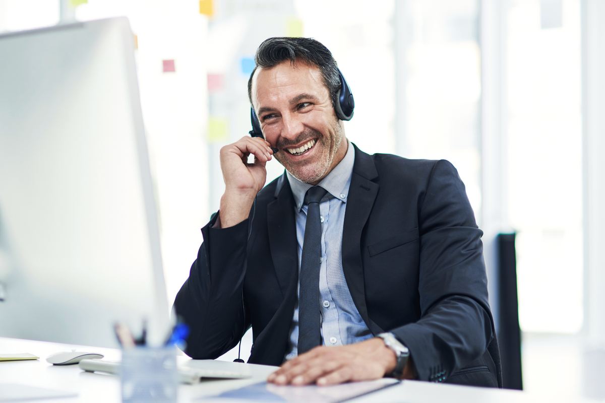 Man in a suit smiling, wearing a headset, sitting at a desk with a computer. Bright office background, suggesting a positive mood.